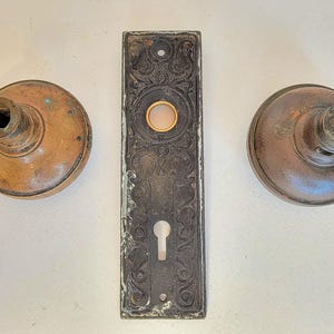 K&ouml;nnte beinhalten: Antikes T&uuml;rbeschlagsset mit zwei runden T&uuml;rkn&ouml;pfen aus Bronze und einer rechteckigen R&uuml;ckplatte. Die R&uuml;ckplatte hat ein verziertes, graviertes Design mit Schl&uuml;sselloch und einer kreisf&ouml;rmigen &Ouml;ffnung. Die T&uuml;rkn&ouml;pfe und die R&uuml;ckplatte weisen Alters- und Gebrauchsspuren auf.