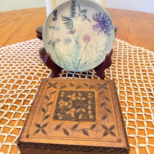 May include: A round ceramic plate featuring a painted hummingbird and purple flowers, displayed on a dark brown stand. In front sits a square wooden box with a dark brown finish, decorated with a carved leaf border and a central floral motif.