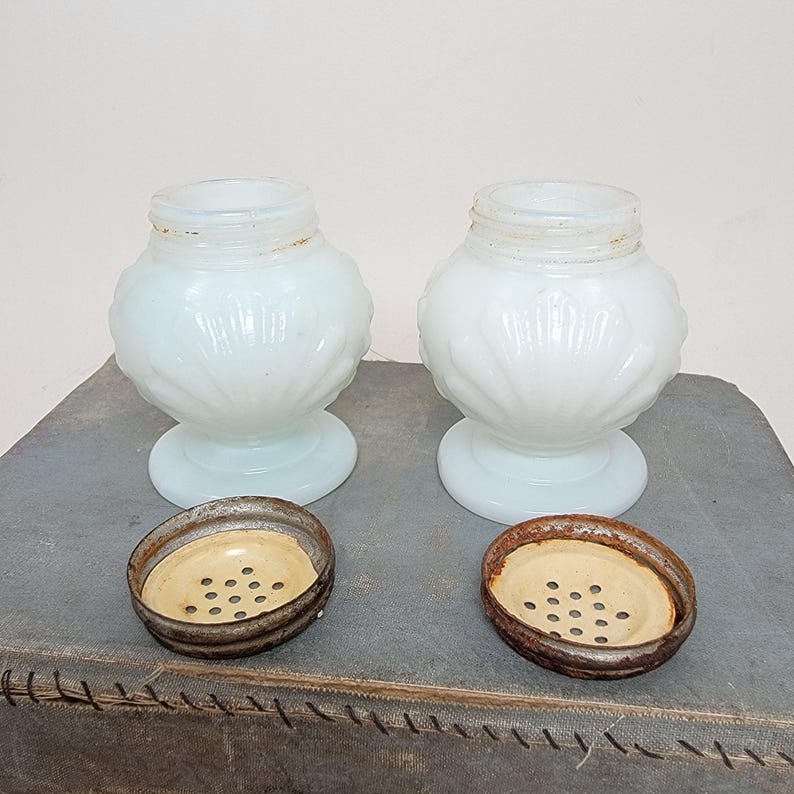 May include: Two antique white glass salt and pepper shakers with decorative floral patterns. The shakers have a bulbous shape and sit on a round base. The metal lids are rusty and have small holes.