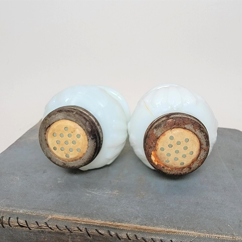May include: Two antique, light blue glass salt shakers with metal tops. The shakers have a ribbed design and a vintage aesthetic. The metal tops have small holes for dispensing salt.
