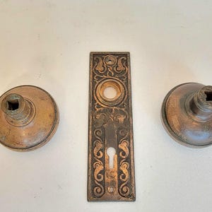 K&ouml;nnte beinhalten: Antikes T&uuml;rbeschlagsset aus Bronze, bestehend aus zwei runden T&uuml;rkn&ouml;pfen und einer rechteckigen R&uuml;ckplatte. Die T&uuml;rkn&ouml;pfe haben quadratische Spindeln. Die R&uuml;ckplatte weist ein verziertes, florales Design mit einem Schl&uuml;sselloch auf.