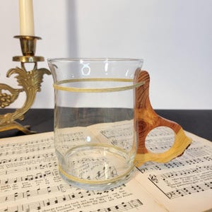 May include: A clear glass drinking cup with a wooden handle. The cup is wrapped with yellow twine. The handle is made of light brown wood. The cup is sitting on top of sheet music. A gold candle holder is in the background.
