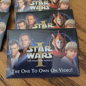 May include: A black and gold Star Wars Episode I: The Phantom Menace magnet featuring a group of characters from the movie. The text "The One To Own On Video!" is printed on the bottom of the magnet.