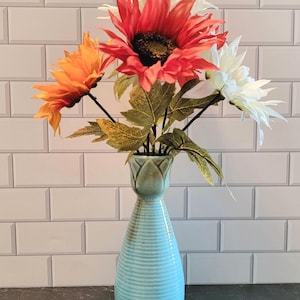 May include: A turquoise ceramic vase holds a bouquet of artificial flowers. The flowers include a large red sunflower, an orange flower, and white flowers. The vase has a ribbed design and a flared base. The background is a white tile wall.