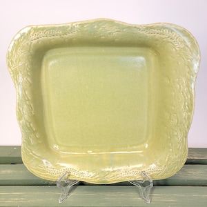 May include: A square, pale green ceramic serving dish with a textured surface and a decorative, scalloped edge. The rim features a detailed leaf and vine pattern. The dish is supported by a clear plastic stand.