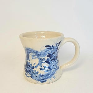 May include: A white ceramic mug with a blue floral design. The mug has a wide base and a narrow top, with a handle on the right side.