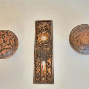 K&ouml;nnte beinhalten: Antikes T&uuml;rbeschlagsset mit zwei runden T&uuml;rkn&ouml;pfen und einer rechteckigen R&uuml;ckplatte. Die T&uuml;rkn&ouml;pfe und die R&uuml;ckplatte aus Bronze weisen aufwendige Blumen- und Rankenmuster auf. Das Set enth&auml;lt ein Schl&uuml;sselloch.