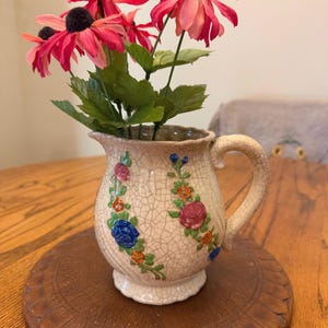 May include: A cream-colored ceramic pitcher with a crackle glaze finish, featuring a handle and embossed floral patterns in blue, pink, and orange. The pitcher is filled with artificial pink flowers with dark centers and green leaves.