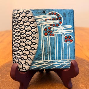 May include: A square ceramic tile featuring a textured silver-grey section with a repeating scale pattern on the left and a bright blue section on the right. The blue area displays white line drawings of three tall trees with red circular accents.