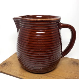 May include: A brown ceramic pitcher with a ribbed design. The pitcher has a wide mouth and a handle.