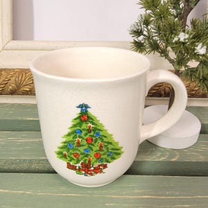 May include: A cream-colored ceramic mug with a crackled glaze finish, featuring a colorful graphic of a decorated Christmas tree with red and blue ornaments, a blue star topper, and red gift boxes at the base, complete with a sturdy side handle.