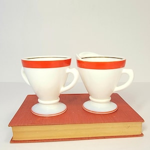 May include: A white creamer and sugar bowl set with a red band and gold trim. The sugar bowl has two handles. The set is on a red book.