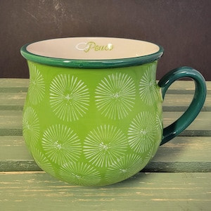 May include: A green ceramic mug with a white floral pattern and a green handle. The word "Peace" is written in cursive inside the mug. The mug has a green rim and a cream-colored interior.