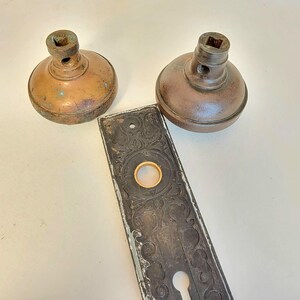 K&ouml;nnte beinhalten: Zwei antike T&uuml;rkn&ouml;pfe aus Bronze und eine dekorative, rechteckige T&uuml;rplatte. Die T&uuml;rkn&ouml;pfe haben eine abgerundete Form mit einem quadratischen Schaft. Die T&uuml;rplatte ist dunkel mit aufwendigen Details und einem goldfarbenen Ring.