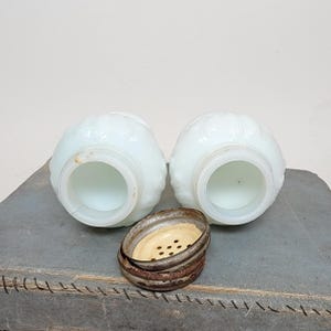 May include: Two antique, white, glass salt shakers with a floral design. The shakers have a round shape and a wide opening. A rusty metal lid with small holes sits between the shakers. The shakers are on a gray surface.