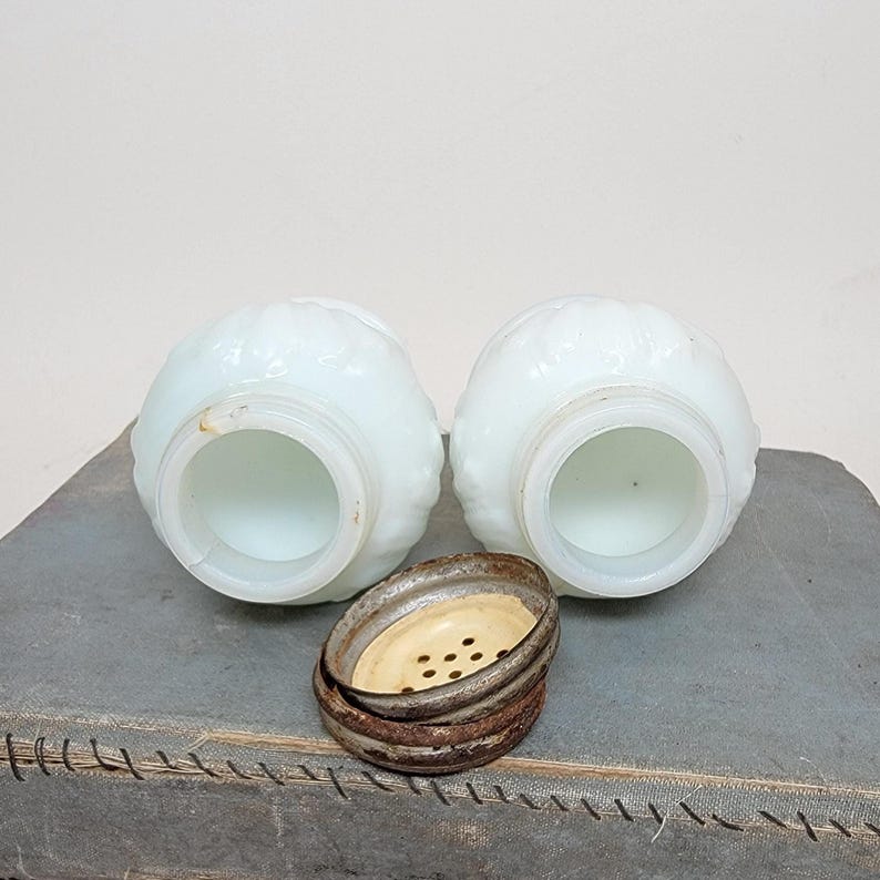 May include: Two antique, white, glass salt shakers with a floral design. The shakers have a round shape and a wide opening. A rusty metal lid with small holes sits between the shakers. The shakers are on a gray surface.