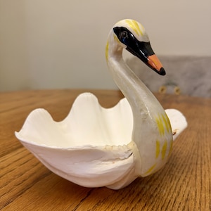 May include: A white swan-shaped decorative bowl with a curved neck and head. The swan features a black beak with an orange tip, black eye markings, and yellow painted accents on the head and neck. The bowl section resembles folded wings.