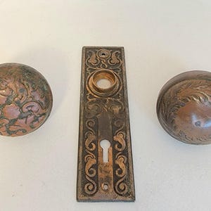 K&ouml;nnte beinhalten: Antikes T&uuml;rbeschlagsset aus Bronze mit zwei runden T&uuml;rkn&ouml;pfen mit floralen Mustern und einer rechteckigen R&uuml;ckplatte mit Schl&uuml;sselloch. Die Beschl&auml;ge weisen Gebrauchsspuren auf.