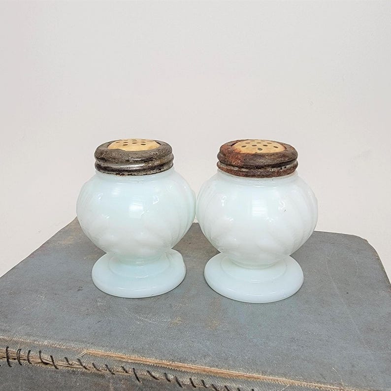 May include: A pair of antique salt and pepper shakers. The shakers are a light blue-white color with a round body and a pedestal base. The lids are a dark metal with small holes for dispensing. The shakers are displayed on a gray surface.