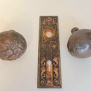 May include: An antique door hardware set featuring two ornate doorknobs and a rectangular escutcheon plate. The doorknobs have a round shape with embossed floral designs. The escutcheon plate has a keyhole.