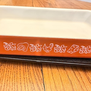 May include: A rectangular baking dish with a bright orange exterior and a smooth white interior. The side of the dish features a repeating white graphic pattern consisting of stylized leaves, a mushroom, a chicken, and a wedge of Swiss cheese.