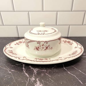 May include: A white ceramic sugar bowl with a lid, resting on a matching plate. Both pieces feature a maroon floral pattern and decorative trim. The set is designed for serving sugar or other condiments.