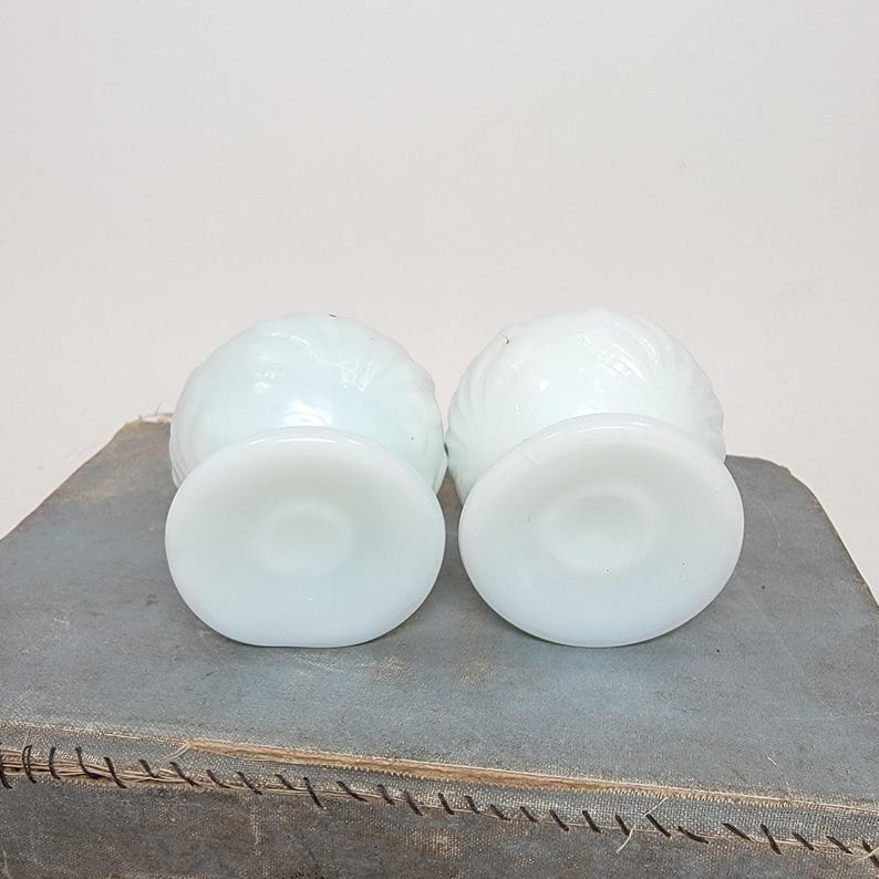 May include: Two white, vintage, swirl-patterned glass dessert dishes. Each dish has a round base and a bowl-shaped top. The dishes are displayed on a gray surface.