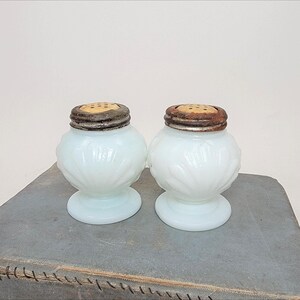 May include: A pair of vintage white glass salt and pepper shakers. Each shaker has a round body with a floral design and a metal lid with small holes. The shakers are displayed on a gray surface.