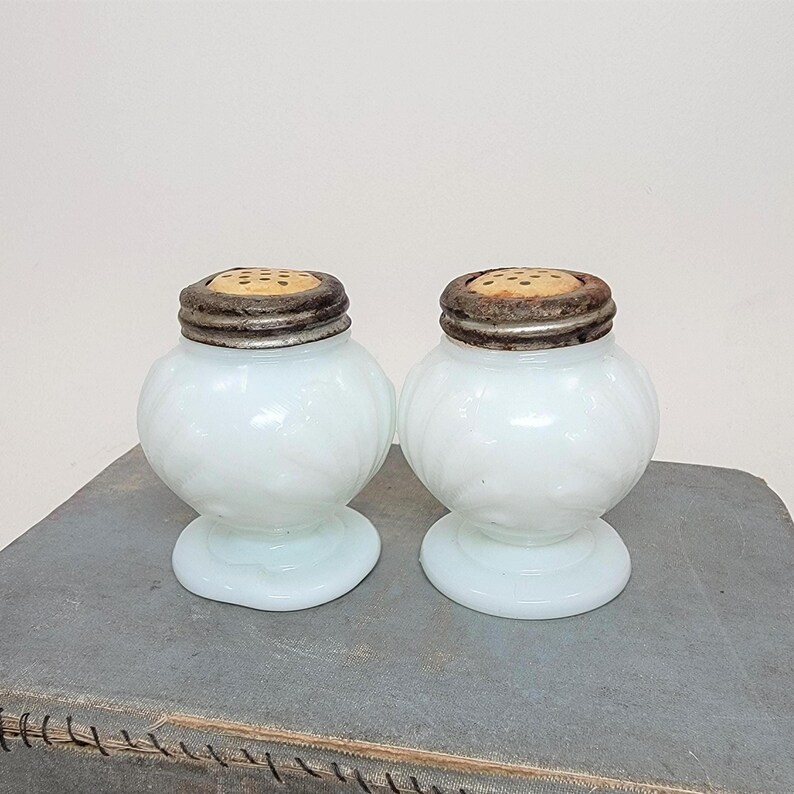 May include: Two antique, opalescent glass salt and pepper shakers. Each shaker has a round body, a pedestal base, and a metal lid with small holes. The glass has a soft, milky white color. The shakers are displayed on a gray surface.