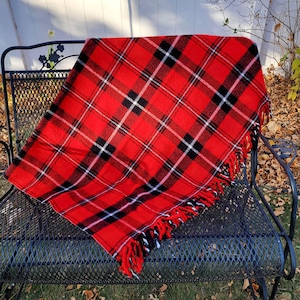 May include: A red plaid blanket with black and white accents and fringe. The blanket is draped over a black metal bench. The plaid pattern features thick black lines and thinner white lines against a bright red background.