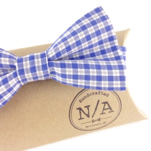 Op de afbeelding: Een blauw en wit geruite vlinderdas. De vlinderdas ligt op een bruine doos met de tekst "Handcrafted N/A Montreal, Qc". De vlinderdas is gemaakt van stof en heeft een klassieke vorm.