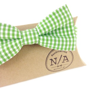 Può includere: Un papillon a quadretti verdi e bianchi appoggiato su una scatola di carta marrone. Il papillon è in tessuto e ha una forma classica. La scatola ha un timbro circolare con la scritta "Handcrafted N/A Montreal, Qc."