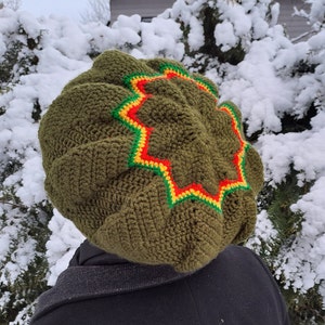 May include: A green crocheted beanie with a yellow, red and green star design. The beanie is being worn by a person standing in front of a snowy background.