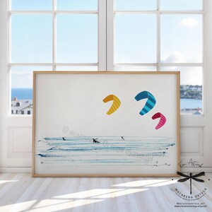 May include: A framed art print featuring a watercolor illustration of kite surfers on the ocean. The artwork showcases three colorful kites in yellow, blue, and pink, set against a blue and white background. The print is displayed in a light brown wooden frame.