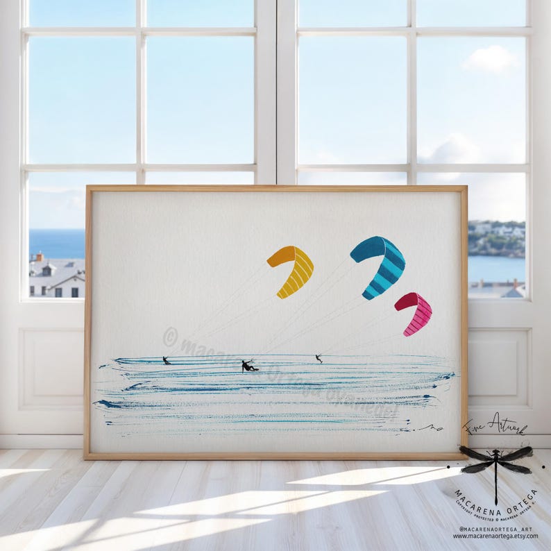 May include: A framed art print featuring a watercolor illustration of kite surfers on the ocean. The artwork showcases three colorful kites in yellow, blue, and pink, set against a blue and white background. The print is displayed in a light brown wooden frame.