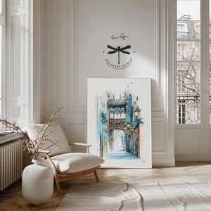 May include: A framed watercolor print of a European street scene with a bridge and archway, rendered in shades of blue, teal, and brown. The artwork is displayed in a bright room with a white chair, a vase with branches, and a rug. The artist's name and logo are visible.