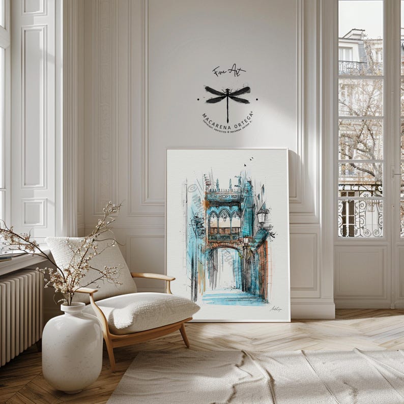 May include: A framed watercolor print of a European street scene with a bridge and archway, rendered in shades of blue, teal, and brown. The artwork is displayed in a bright room with a white chair, a vase with branches, and a rug. The artist's name and logo are visible.