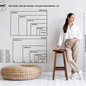 May include: A size guide chart in black displays dimensions in centimeters for European and international frames, with corresponding inch measurements. A woman stands near a wooden stool, with a woven ottoman in the foreground. The height is 66.14 inches (168 cm).