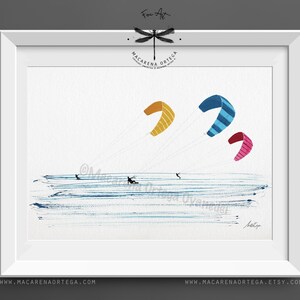 May include: Watercolor painting of kite surfers on the water with colorful kites. The kites are yellow, blue with stripes, and pink with stripes. The artwork is framed in a white frame. The artist's name and website are visible.