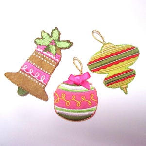 May include: Three felt Christmas decorations: a bell with a green holly sprig, a striped ornament with a green, red, and yellow design, and a pink and green striped bauble with a pink bow.
