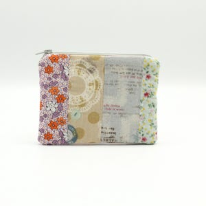 Op de afbeelding: Een kleine, rechthoekige ritszak met een patchwork-ontwerp. De tas is gemaakt van verschillende stoffen met bloemenpatronen in roze, oranje en witte tinten. De stof heeft ook een gedrukte tekst in zwart.