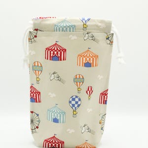 May include: A beige drawstring bag with a white cord. The bag has a pattern of red and white striped tents, blue and white checkered hot air balloons, and cartoon birds.