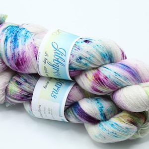 May include: Two skeins of white yarn featuring a speckled pattern of vibrant blue, purple, and lime green. Each skein is wrapped in a paper label with the text Libbyroo Yarns, A hop, stitch and jump away, and other printed information.