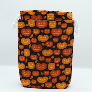 Peut inclure: Un sac à cordon sur le thème d'Halloween. Le sac est noir avec un motif répété de citrouilles orange. Il possède une fermeture à cordon blanche en haut. Le sac est en tissu.