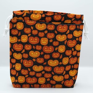 Könnte beinhalten: Ein Halloween-Kordelzugbeutel mit einem sich wiederholenden Muster aus orangefarbenen Kürbissen auf schwarzem Hintergrund. Der Beutel hat einen weißen Kordelzugverschluss und ist aus Stoff.
