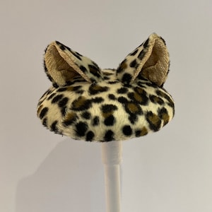 May include: A small, round, leopard print hat with two furry ears. The hat is on a white stand.