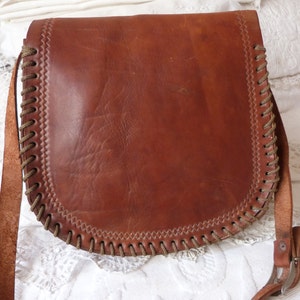 May include: A brown leather shoulder bag with a braided trim and a buckle closure. The bag has a long strap and is made of a soft, supple leather.