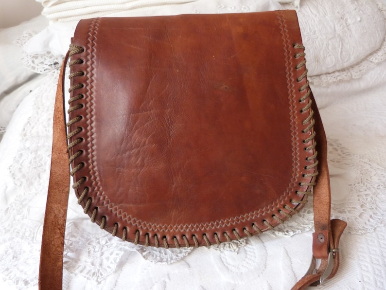 May include: A brown leather shoulder bag with a braided trim and a buckle closure. The bag has a long strap and is made of a soft, supple leather.