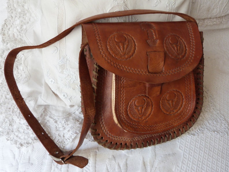 May include: A brown leather shoulder bag with intricate embossed floral designs. The bag has a flap closure and a long adjustable strap.