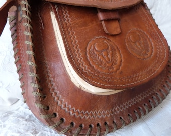 May include: A brown leather satchel bag with a flap closure and a decorative stitched design. The bag has two embossed circular designs with a stylized animal head and leaves.